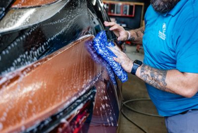 Brian, owner of Summer Breeze Ceramic Coating & Mobile Detailing, wears his company's blue uniform shirt while exterior detailing a car.